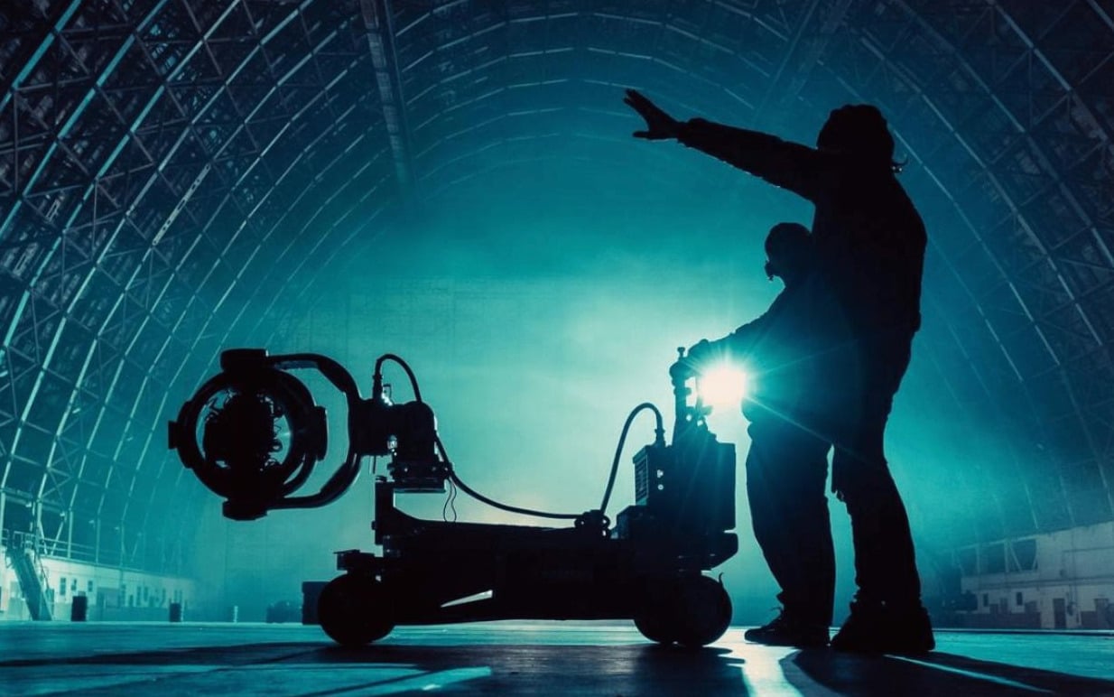 Silhouettes of two people with a camera rig in a large, blue-lit hangar.